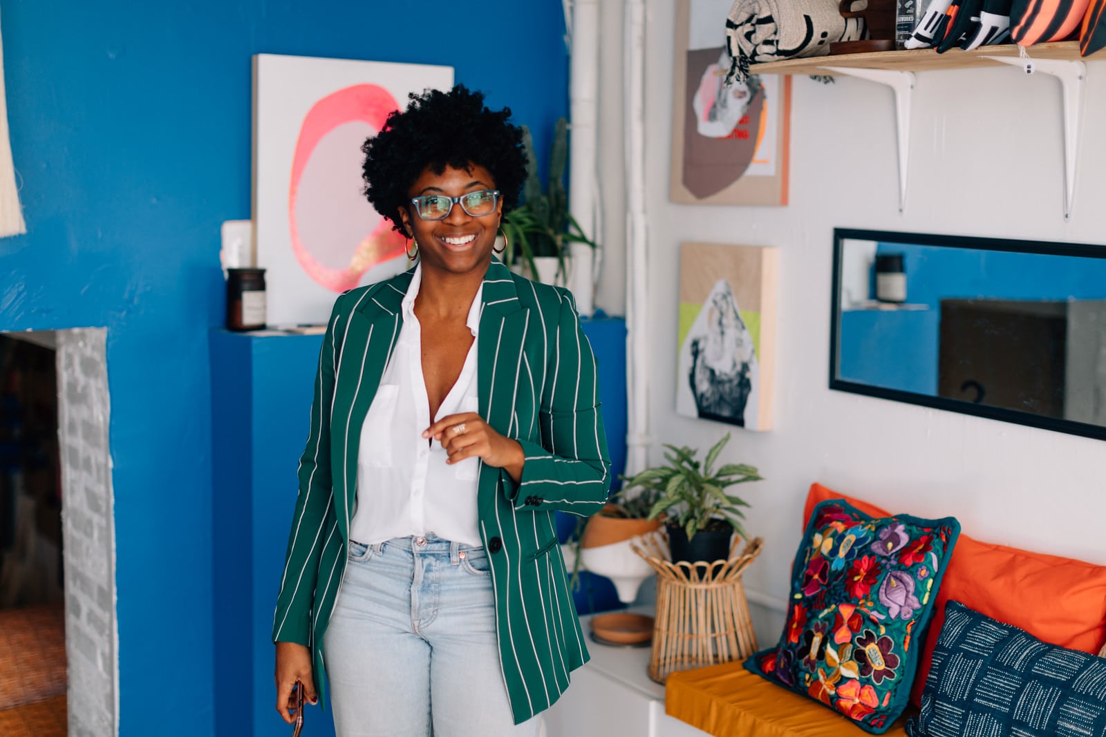 A smiling black woman standing in a confident pose, wearing a white shirt, jeans, and a green and white blazer. She is standing in front of a blue wall and orange couch.